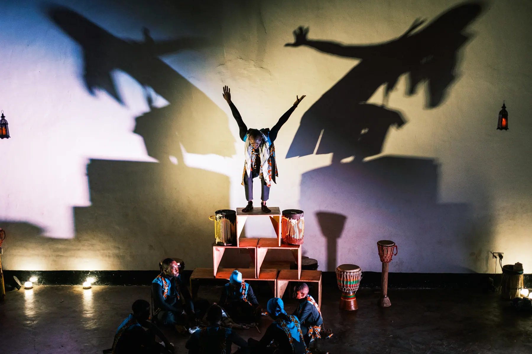 A Sawa Ensemble performer raising both arms as stage lighting casts large shadows
